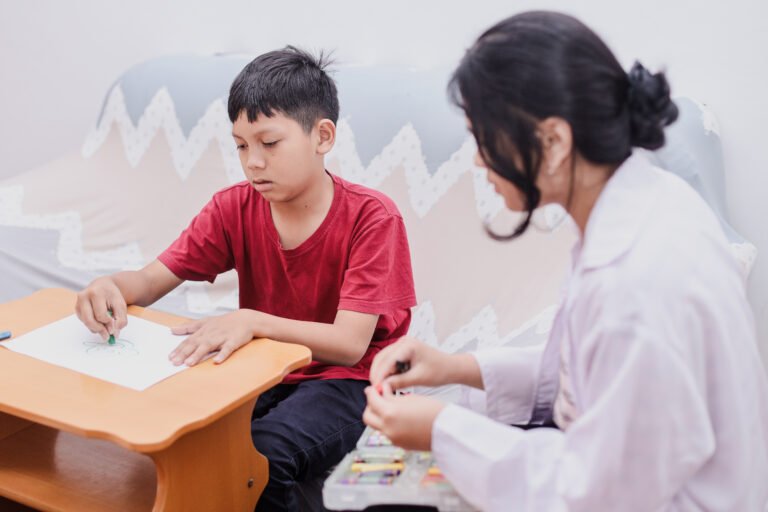Boy drawing at a small table with an adult nearby.