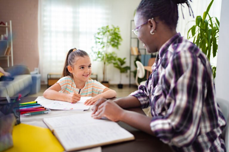 Girl drawing at a table with an adult beside her.