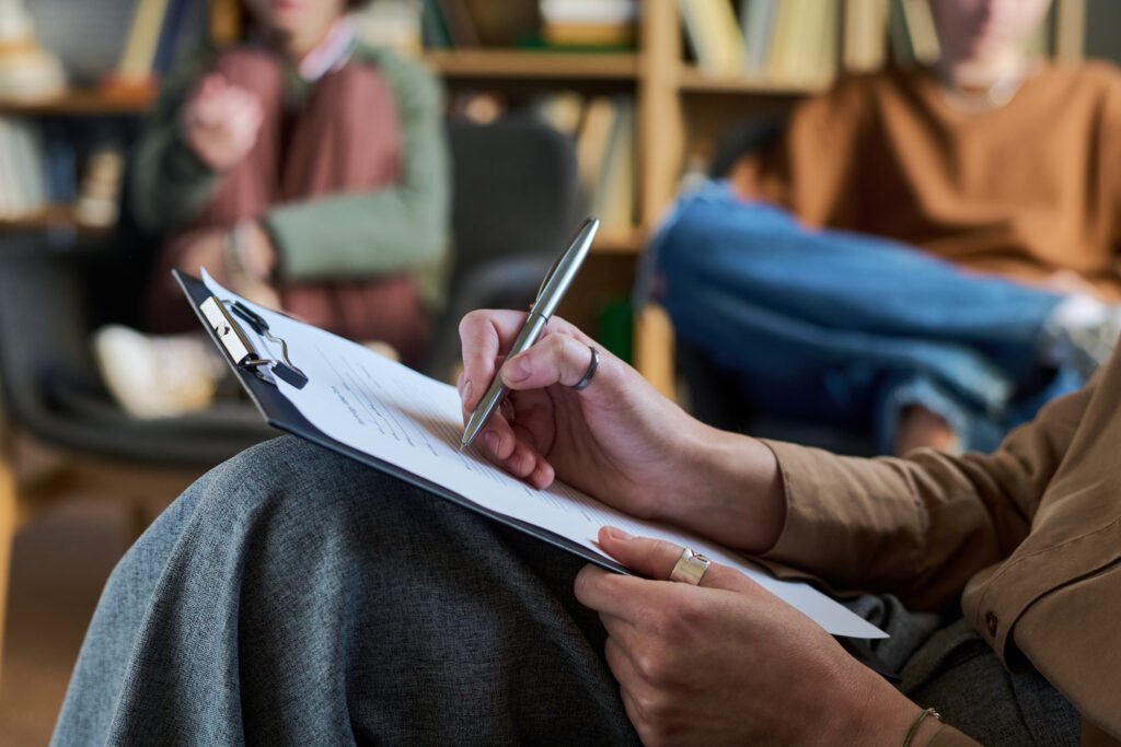 Person writing on a clipboard during a seated discussion.